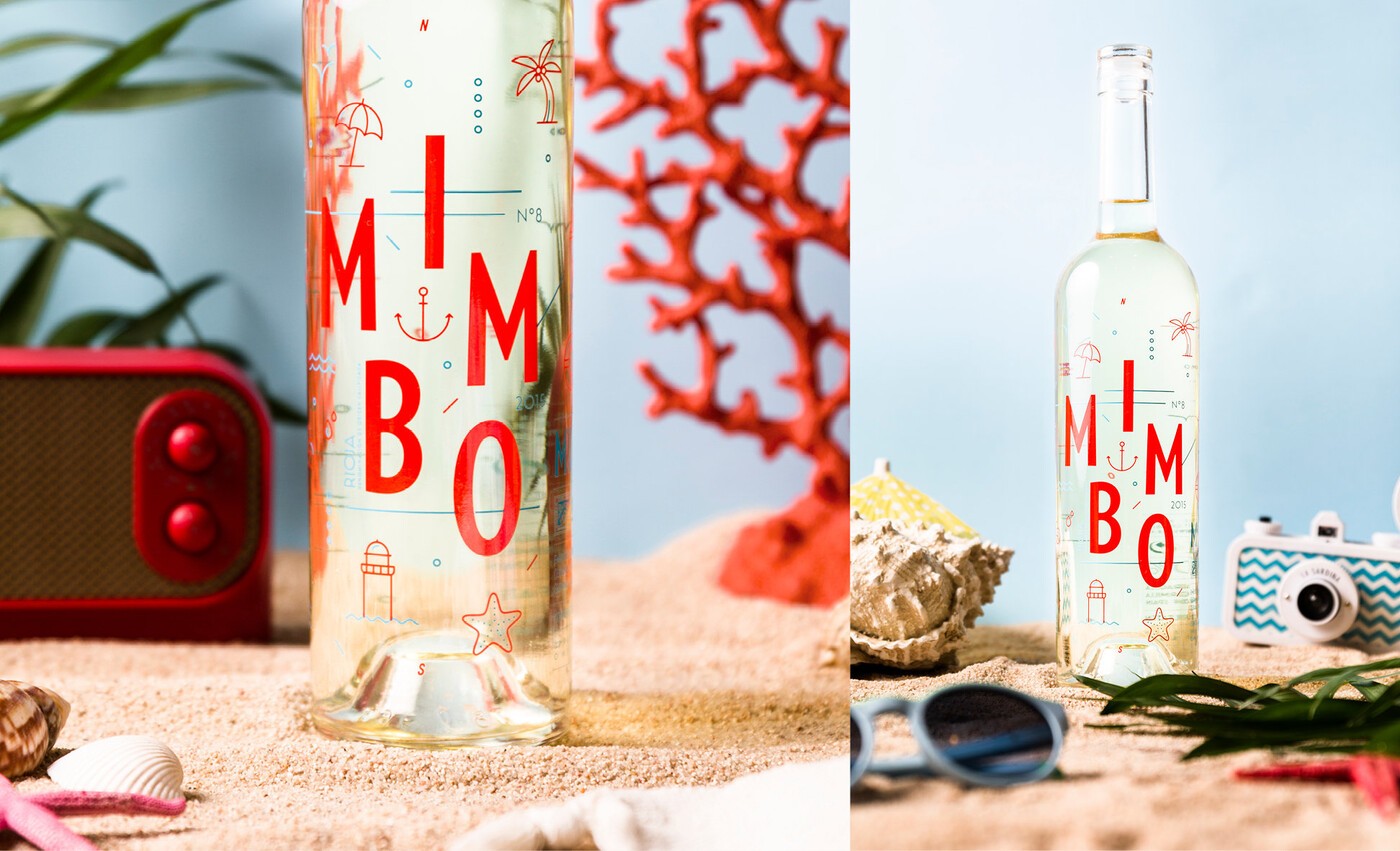 Bodegón ambientado en una mañana de playa, con una botella de Mimbo blanco sobre arena de playa, una radio, coral rojo y una cámara de fotos analógica